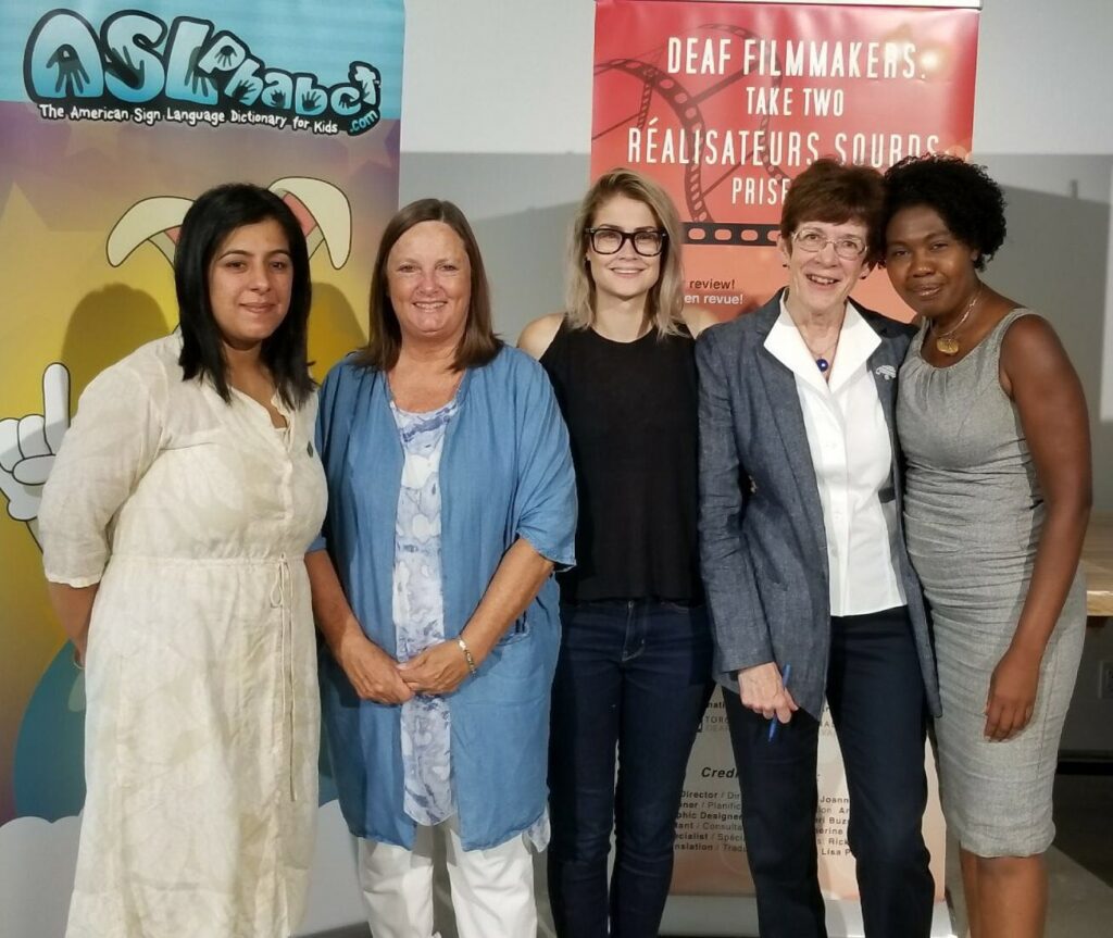 Photograph of five people standing in front of colourful banner signs. They are (from left to right) PA Harinder Malhi, Joanne Cripps (Deaf Culture Centre), Rylyn Lennox (Deaf Culture Centre), Diane Davy (Work in Culture), and Claudia McKoy (UpSurgence).